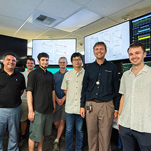 Seven men stand in front of computer screens with data