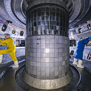 Two scientists stand inside a vacuum chamber with a large cylindrical post in the center