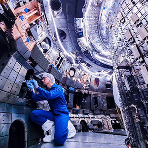 An engineer in a blue jumpsuit uses tools in a narrow opening inside a vacuum vessel