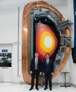 Two men stand in front of the DIII-D tokamak cross-section display