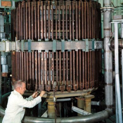 Man in white lab coat working on cylindrical vacuum chamber surrounded with copper coils